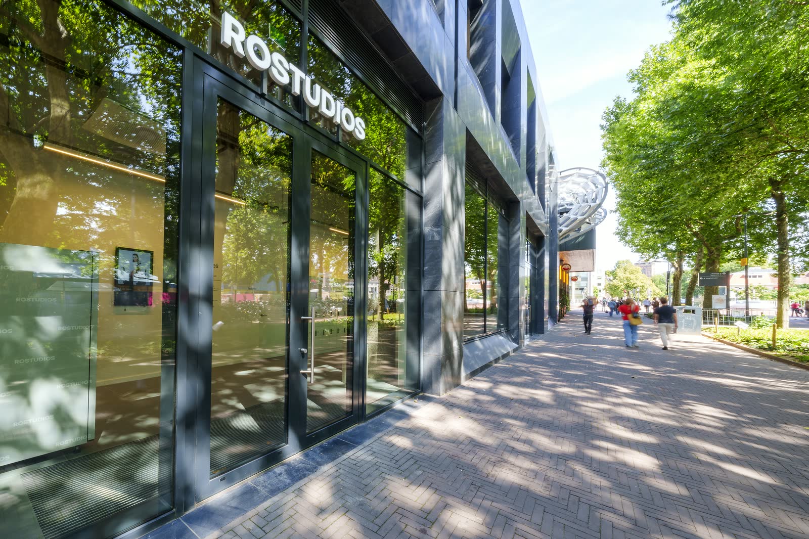 Glazed facade with ROSTUDIOS lettering above the entrance, on a tree-lined street with brick paving