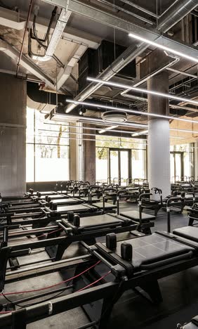 Pilates studio with rows of reformers, concrete columns, exposed ceiling services and daylight from full-height windows