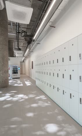 Corridor with a long wall of pale grey lockers, exposed ceiling services and pipework, and dappled sunlight across the tiled floor