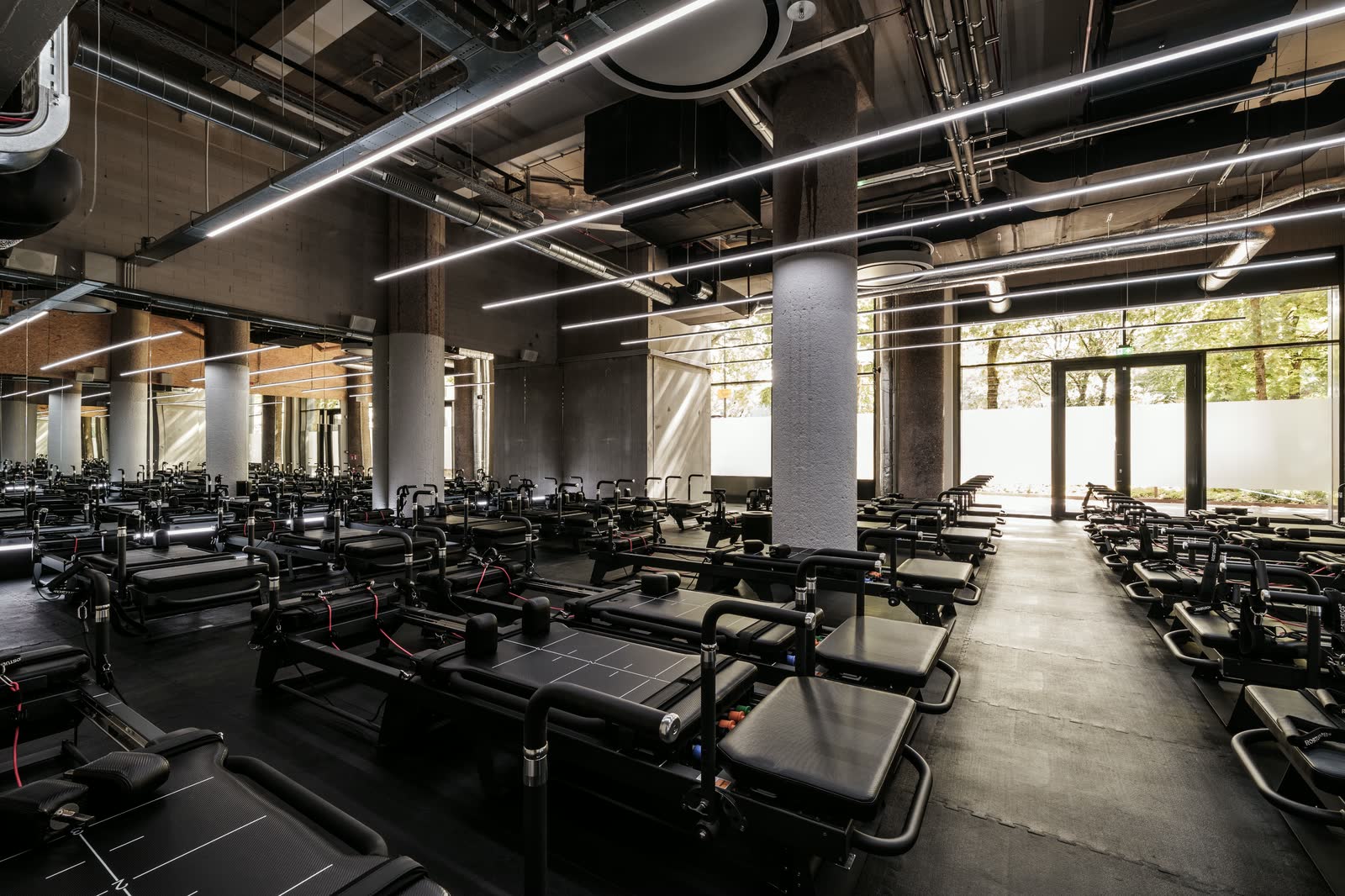 Pilates studio with rows of reformers, linear ceiling lights, a mirrored wall and a glazed entrance to the garden