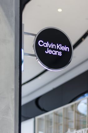 Illuminated circular blade sign reading Calvin Klein Jeans, projecting from the shopfront inside a shopping centre