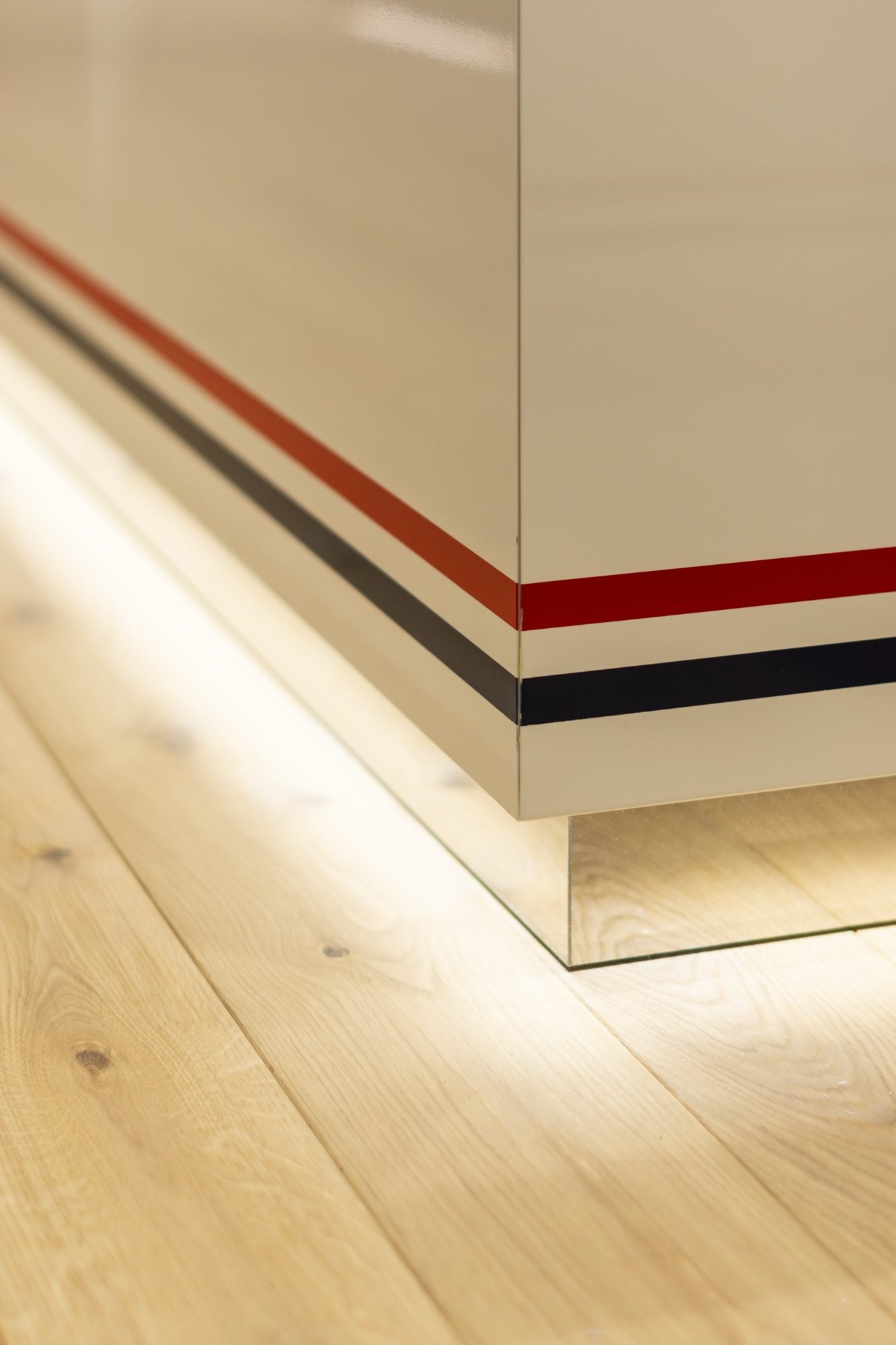 Detail of a display unit edge with red and navy banding above an illuminated recess and oak floor