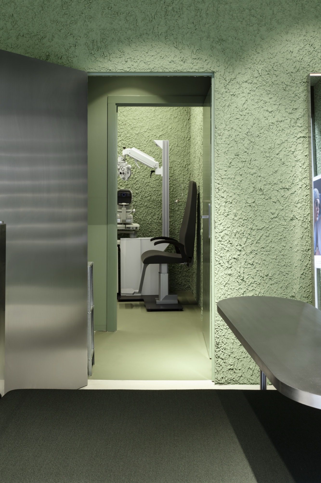 Eye examination room with green textured plaster walls, a slit lamp and phoropter on a white cabinet, seen through an open door