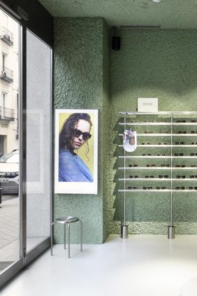 Store corner with green textured plaster walls, brushed-steel eyewear display rails, a framed portrait photograph and a metal stool beside the glazed shopfront