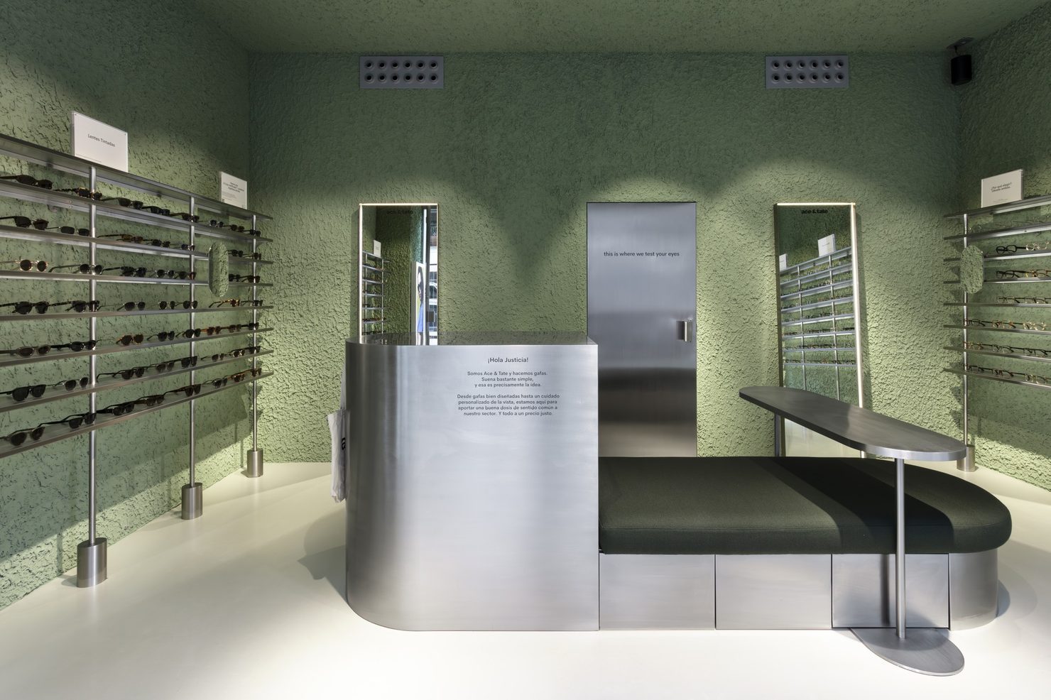Store interior with green textured plaster walls, a curved brushed-steel counter and bench seat, two full-height mirrors and eyewear rails on three walls