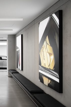 Showroom corridor with large backlit brand images in black frames on concrete-look wall panels, low dark bench seating below and a car visible beyond