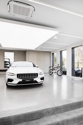 Showroom with a white car beneath a suspended luminous ceiling panel, a motorcycle alongside and full-height glazing onto greenery